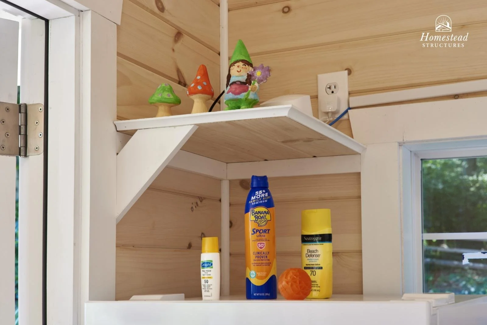 A small shelf with decorative mushroom and gnome figurines above bottles of sunscreen and a small orange rubber ball in a corner of a wooden room with a window.