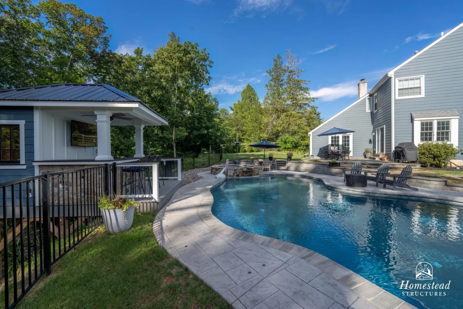 Backyard with a swimming pool, blue house, patio area with seating, surrounded by trees and greenery.