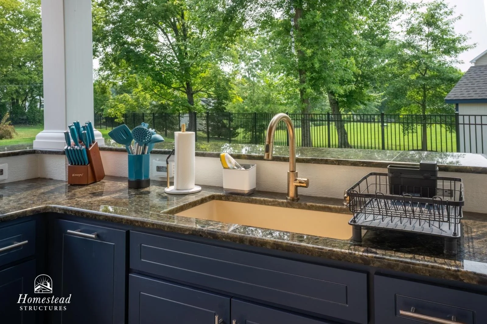 Kitchen counter with blue cabinets, over a granite countertop, with a soap dispenser, dish rack, paper towel holder, and various cooking utensils, overlooking a backyard with trees and a fence.