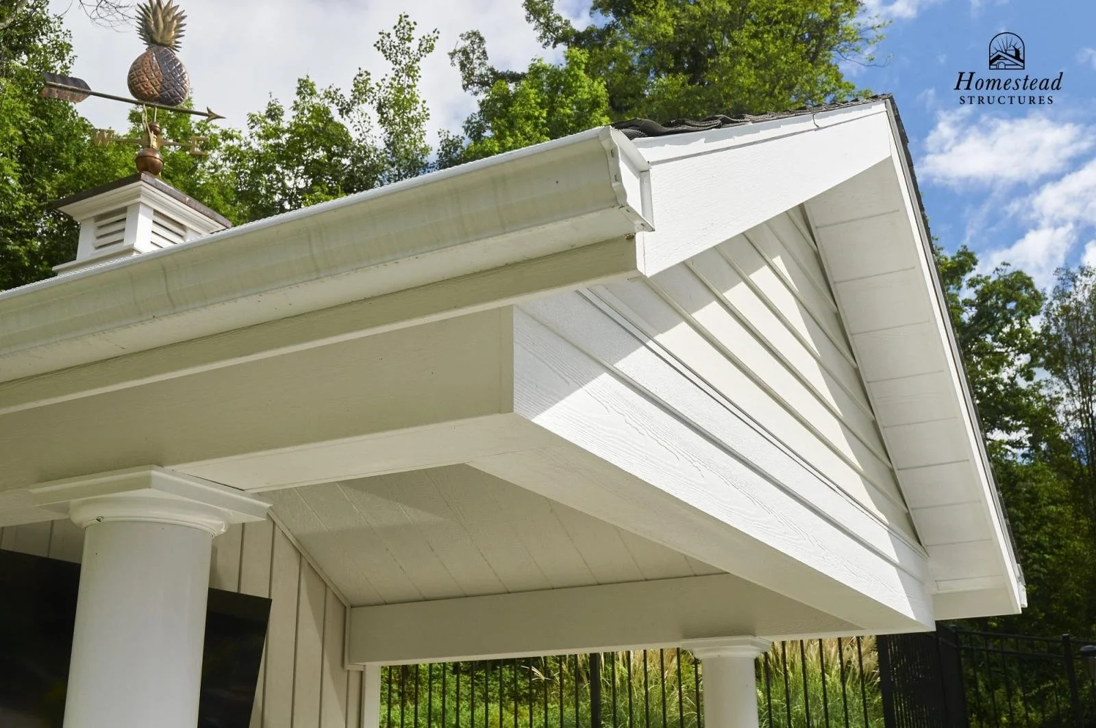 Close-up view of a white house's gable roof with decorative pillars, light-colored siding, and a cupola with a weather vane on top. Green trees and blue sky with clouds in the background.