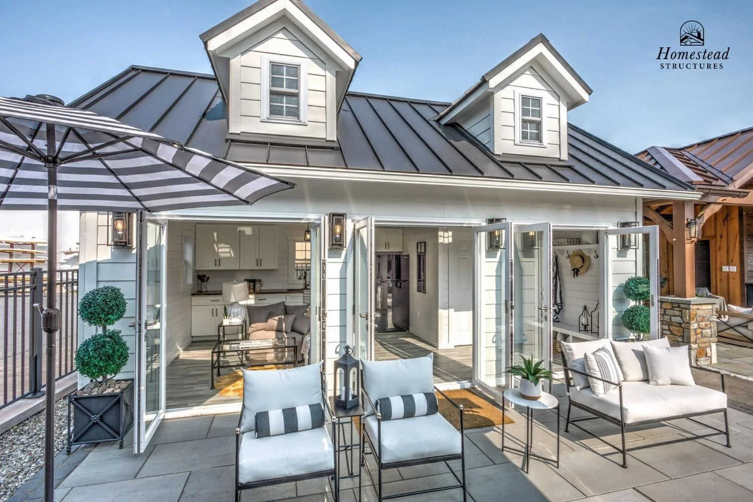 A modern backyard with a tiny house, outdoor seating, and an umbrella. The tiny house has white siding, open glass doors, and a metal roof with dormer windows. The patio features white cushioned chairs, a small table, and decorative plants. The scene