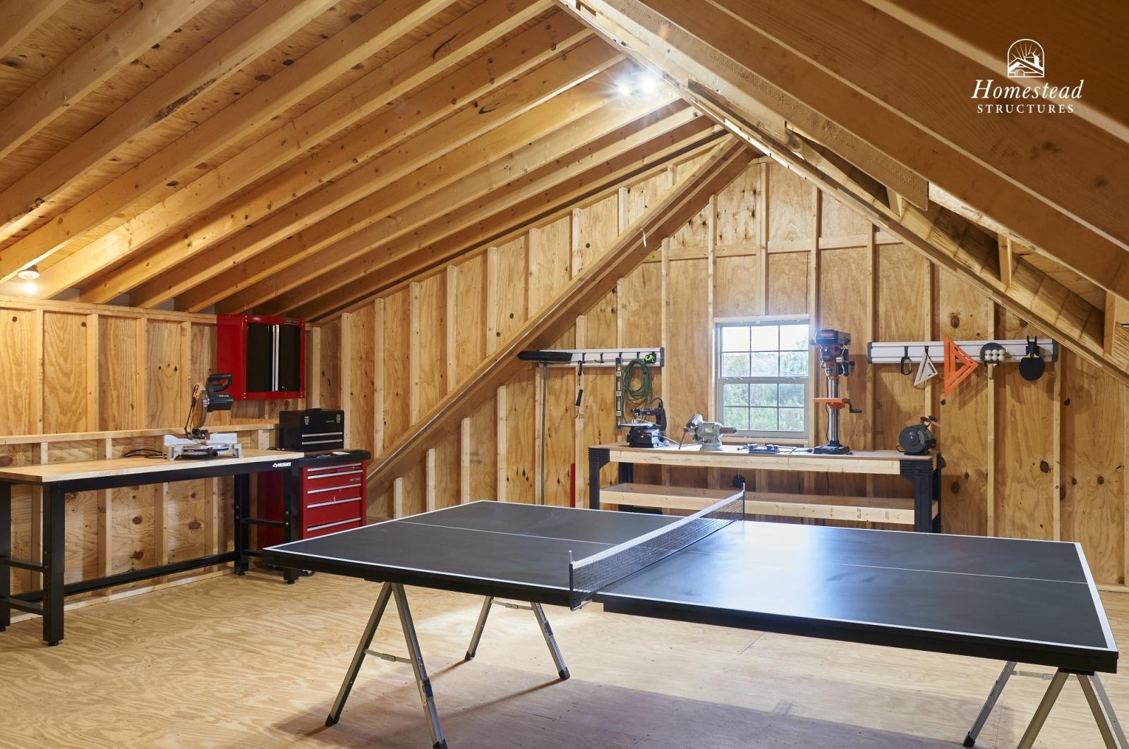 A spacious woodworking garage with a ping pong table in the center, surrounded by workbenches, tools, and storage cabinets, featuring wooden walls and a sloped ceiling with a small window.