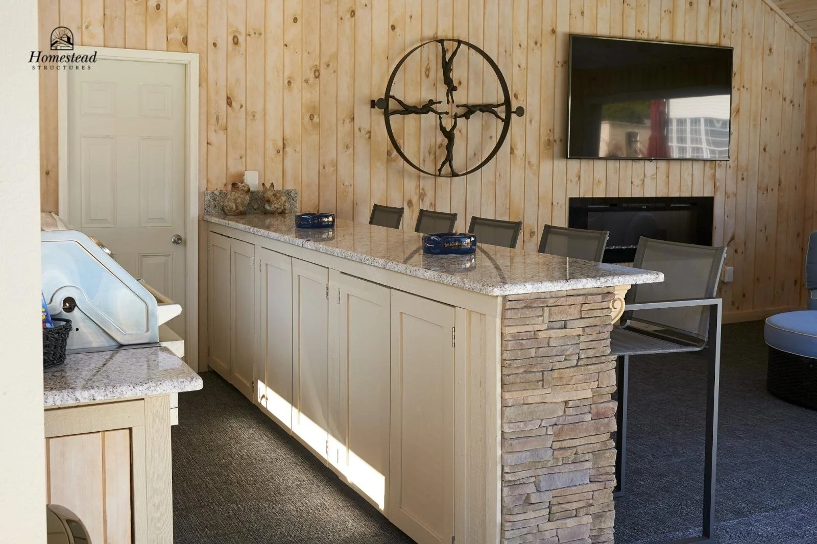 A kitchen area with a long granite countertop, white cabinets, and a stone partial wall. The wall behind the counter is made of wooden planks and features a large circular sculpture with human figures. A flat-screen TV is mounted above a fireplace, a