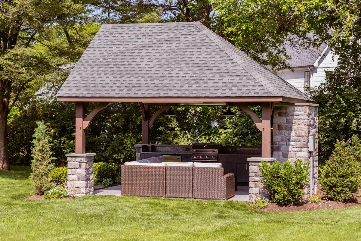 Outdoor pavilion with a stone and wood structure, a pitched roof, outdoor kitchen, grill, and seating area, surrounded by grass and trees.