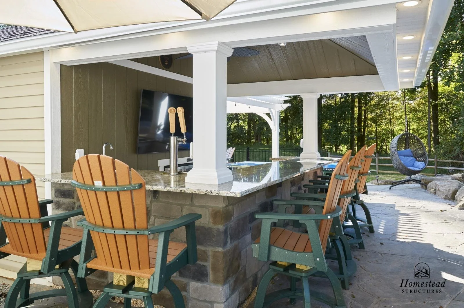 An outdoor covered bar area with a granite countertop, several wooden and green chairs, a mounted flat-screen TV, a hanging swing chair with blue cushions, and a view of trees in the background.