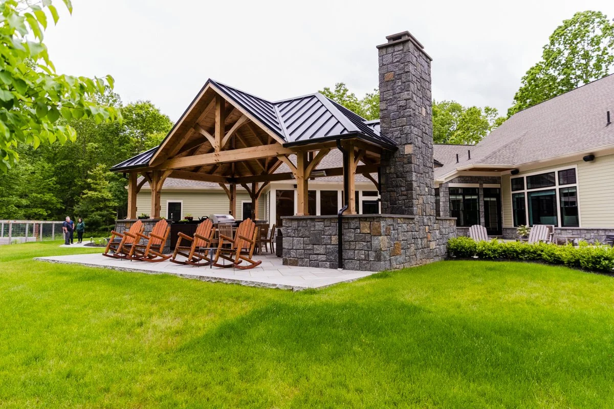 Backyard patio with wooden pergola, stone fireplace, and Adirondack chairs surrounded by green grass and trees.