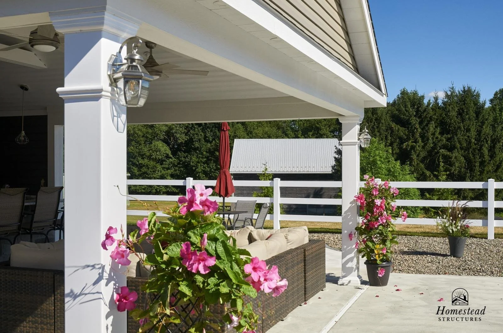 An outdoor patio area with a white column, pink flowering plants in pots, a wicker sofa with beige cushions, and a table with an umbrella. There are trees and a white fence in the background.