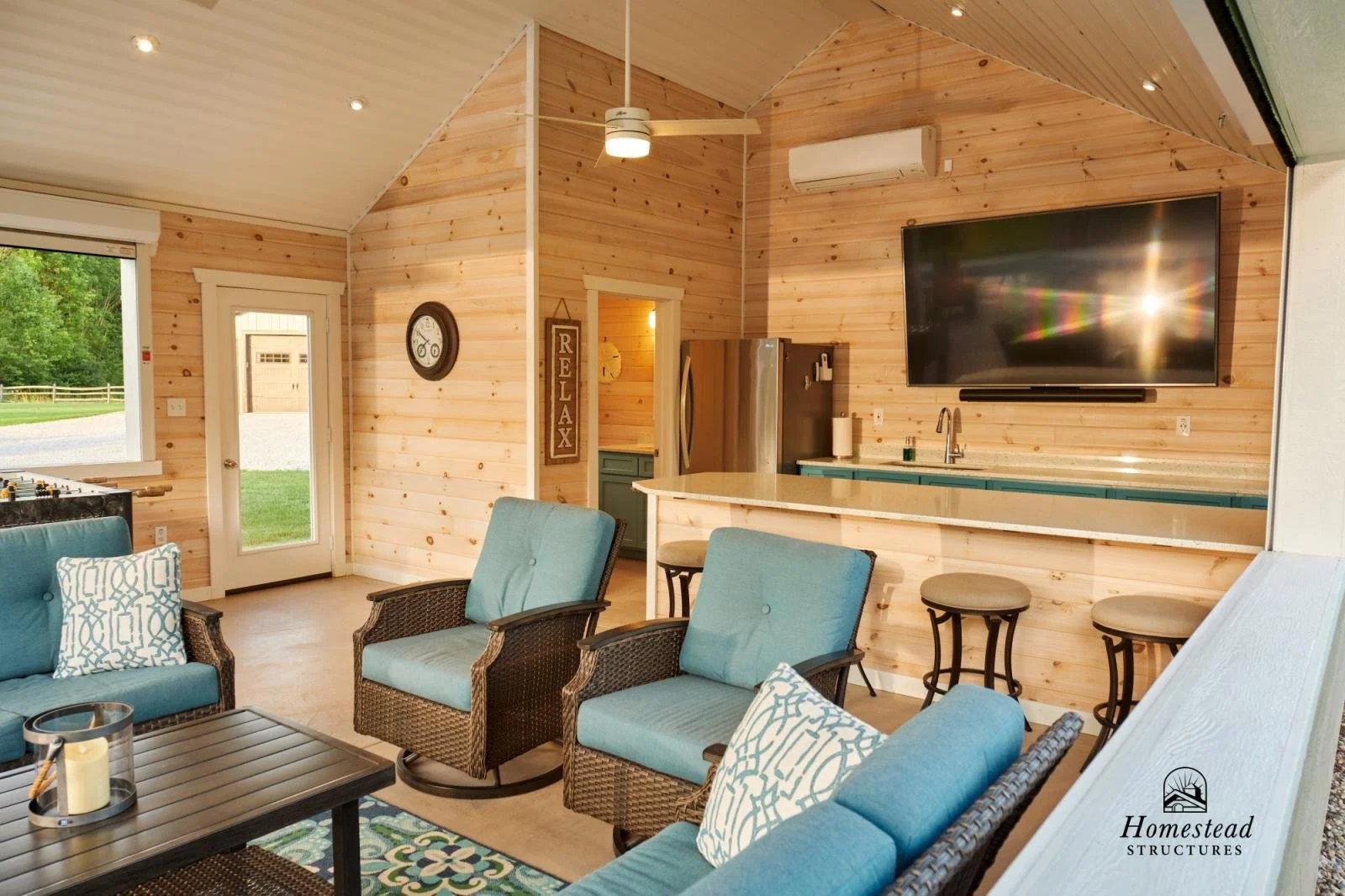 Living area with blue cushioned wicker chairs and matching pillows, wooden walls, a large wall-mounted TV, a kitchen with a stainless steel fridge, bar stools, a small window and door with a view of green trees outside.