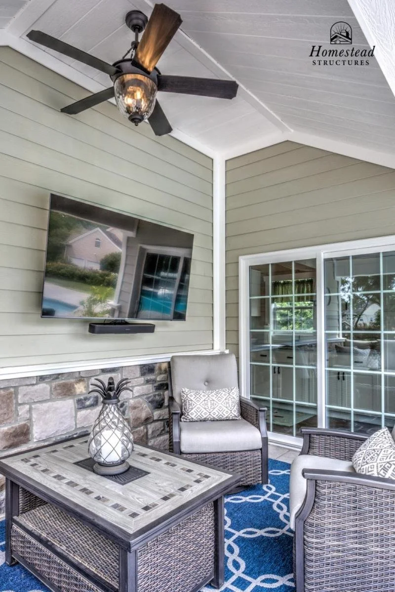 An outdoor covered patio with a ceiling fan, wall-mounted TV, wicker furniture with cushions, a blue patterned rug, a glass door leading inside, and a decorative pineapple lantern on a table.