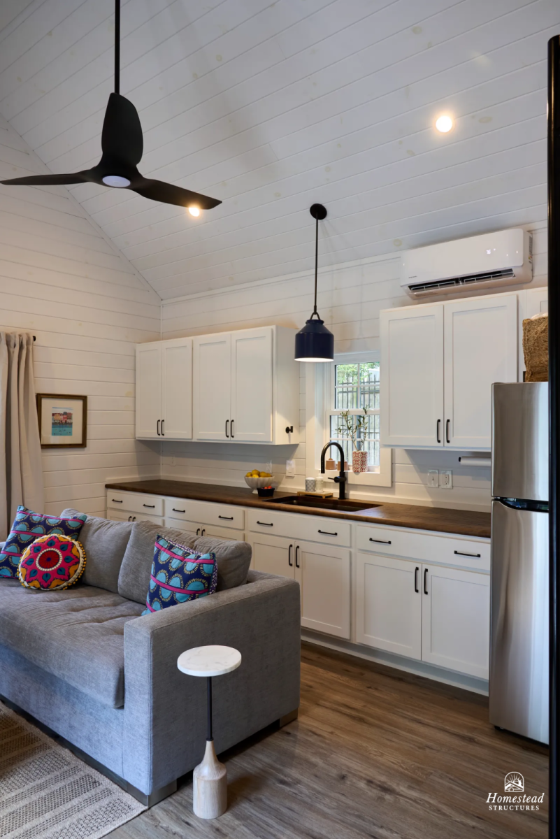 Living room with gray sofa and colorful pillows, open kitchen with white cabinets, wooden countertop, small window, air conditioning unit, ceiling fan, hanging light, wooden flooring.