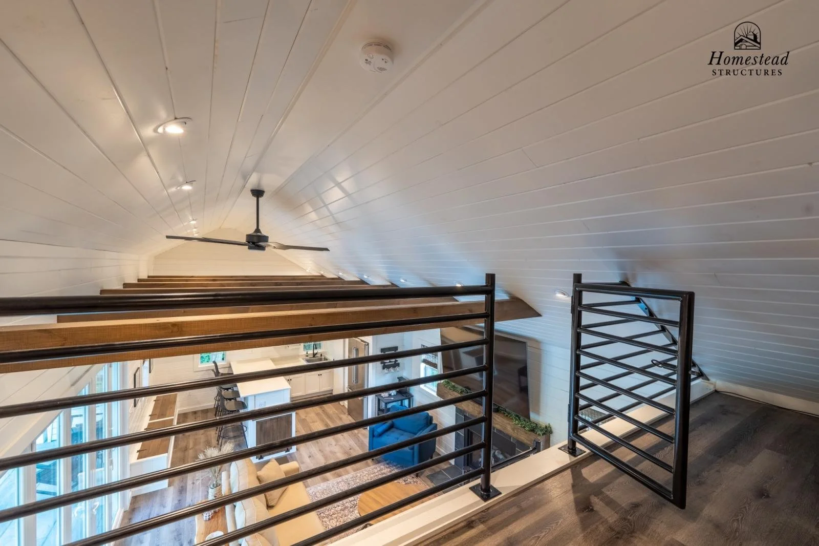 Interior view of a loft with a black ceiling fan, recessed lighting, wood flooring, and a metal railing, overlooking a living area with a couch, TV, fireplace, and large windows.