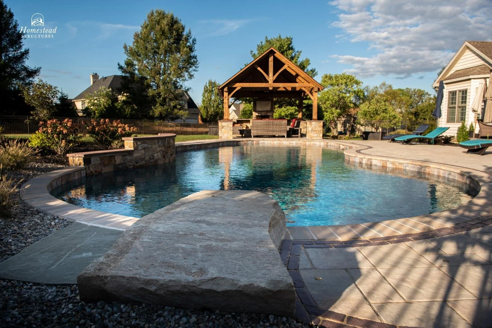 A backyard with a kidney-shaped swimming pool, a shaded sitting area with a stone fireplace, multiple lounge chairs on a concrete patio, and trees in the background.