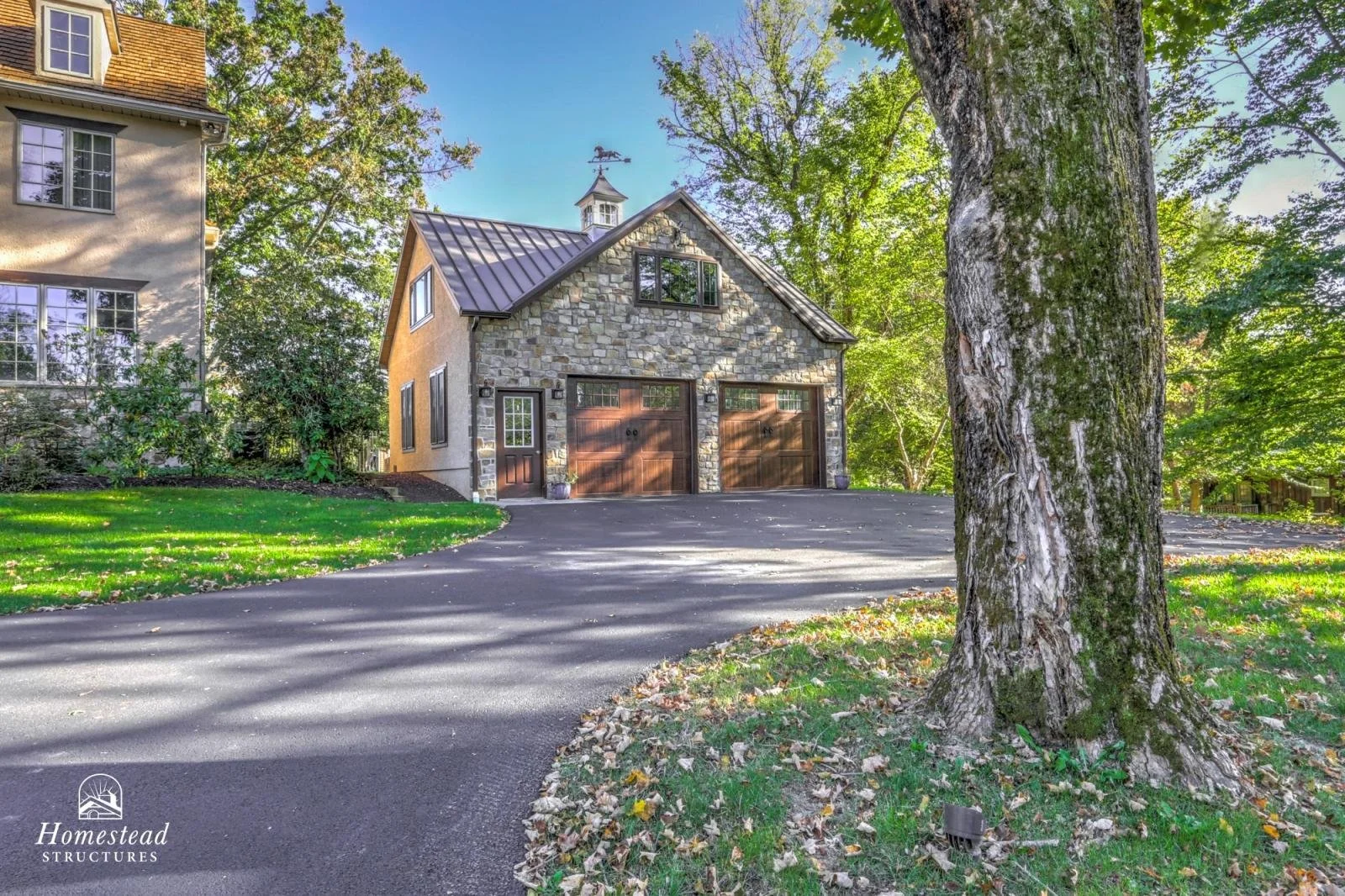 A stone and wood two-car garage with a small door on the left, adjacent to a large tree on a lush green lawn, with another house partially visible to the left, surrounded by trees in a suburban neighborhood.