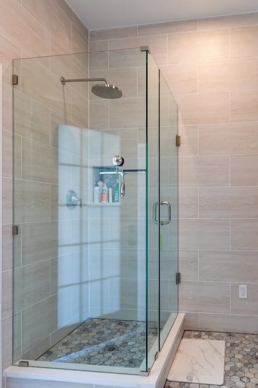 Modern glass shower with beige tiled walls, a rainfall showerhead, and built-in shower shelf with toiletries, in a bathroom with hexagonal floor tiles.