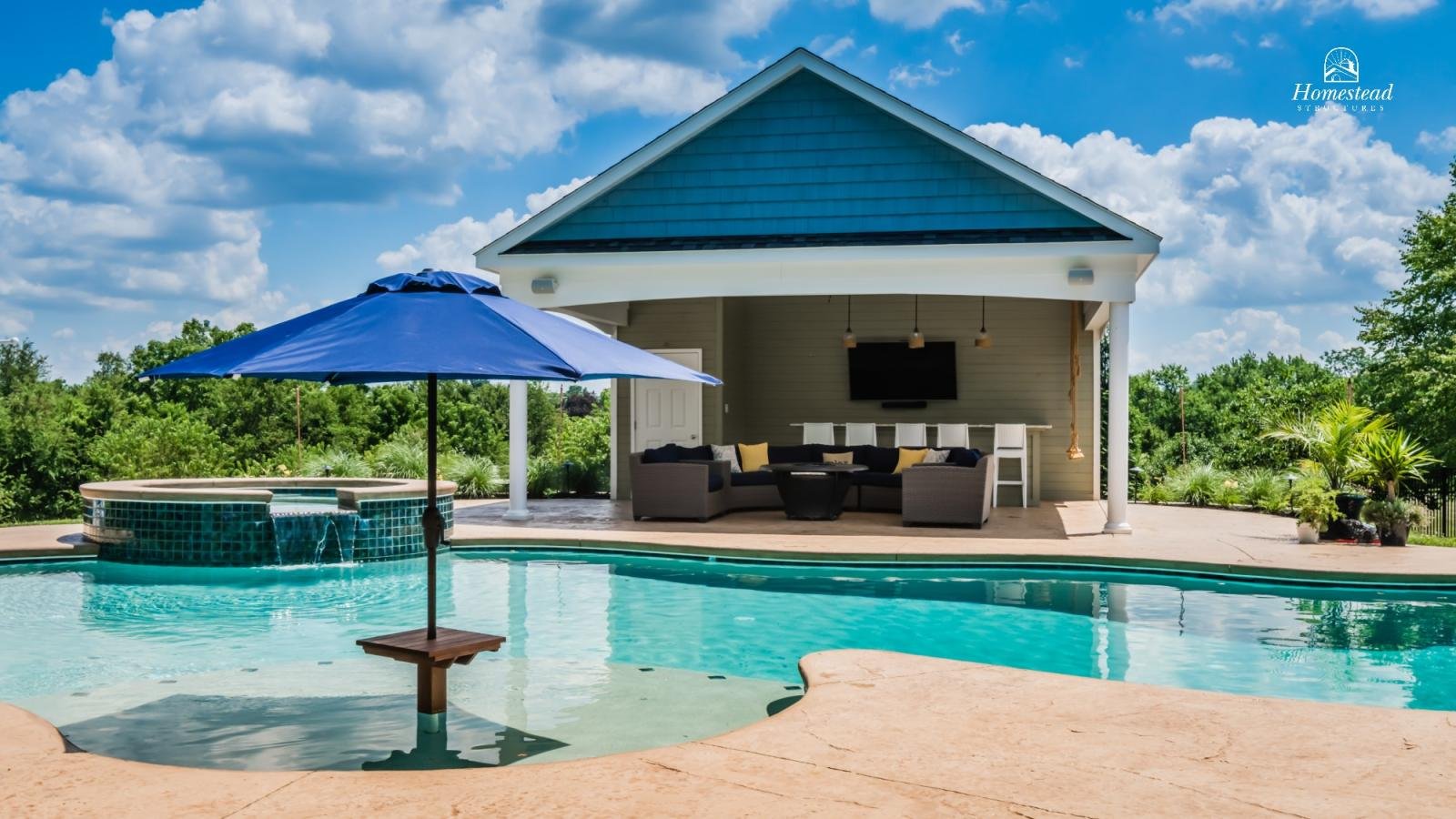 Backyard pool with blue umbrella, hot tub, outdoor seating area, large TV, and lush green trees in the background.