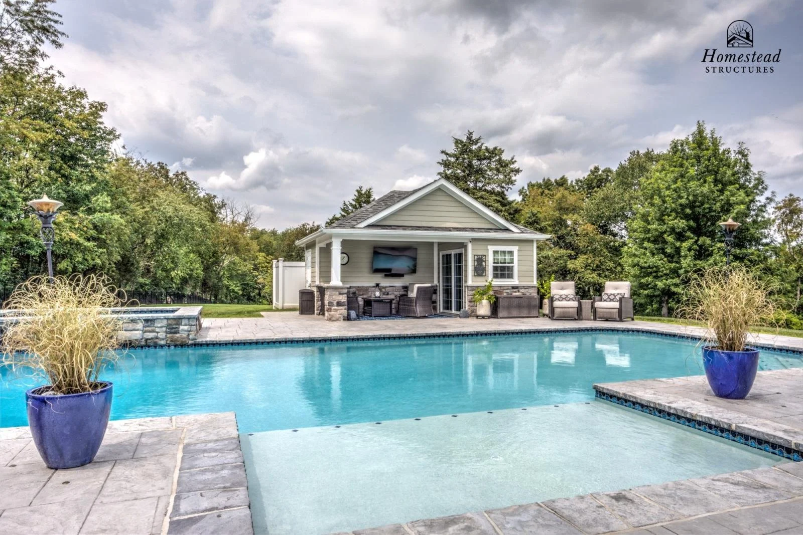 Backyard swimming pool with patio furniture, potted plants, and a small house or pool house in the background, surrounded by trees.