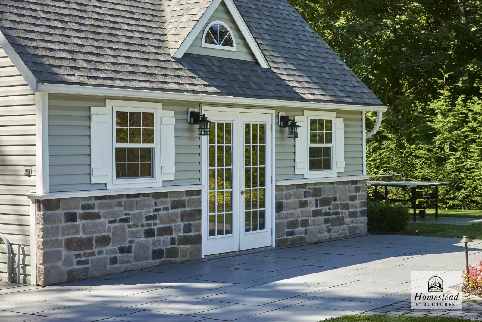 Exterior of a house with siding, stone foundation, and a set of glass French doors. Two wall-mounted lantern lights flank the doors. There are two windows with white shutters on either side of the door. The roof has shingles and a small gable window.