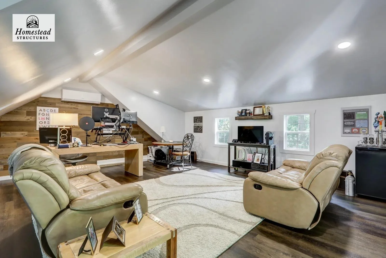 Living room with two beige leather recliners, a wooden table with framed photos, a white area rug, a TV on a black stand, a small desk with a computer, and a wooden accented wall with various decor. There are windows and ceiling lights illuminating t
