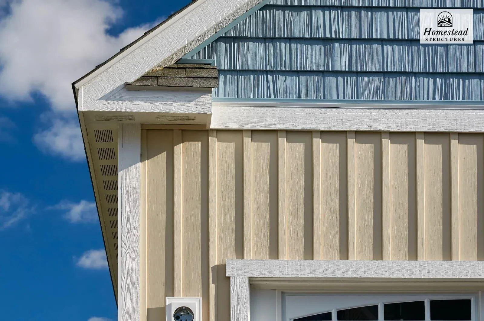 Close-up of the upper corner of a building showing beige vertical siding, white trim, blue decorative siding, and roof with shingles, under a partly cloudy sky.