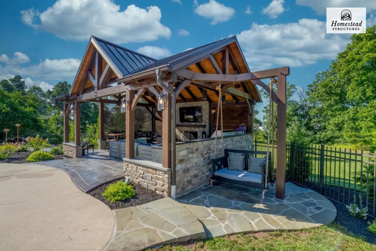 Outdoor living space with a wooden pavilion, swing, stone fireplace, seating area, and lush green landscape under a blue sky.