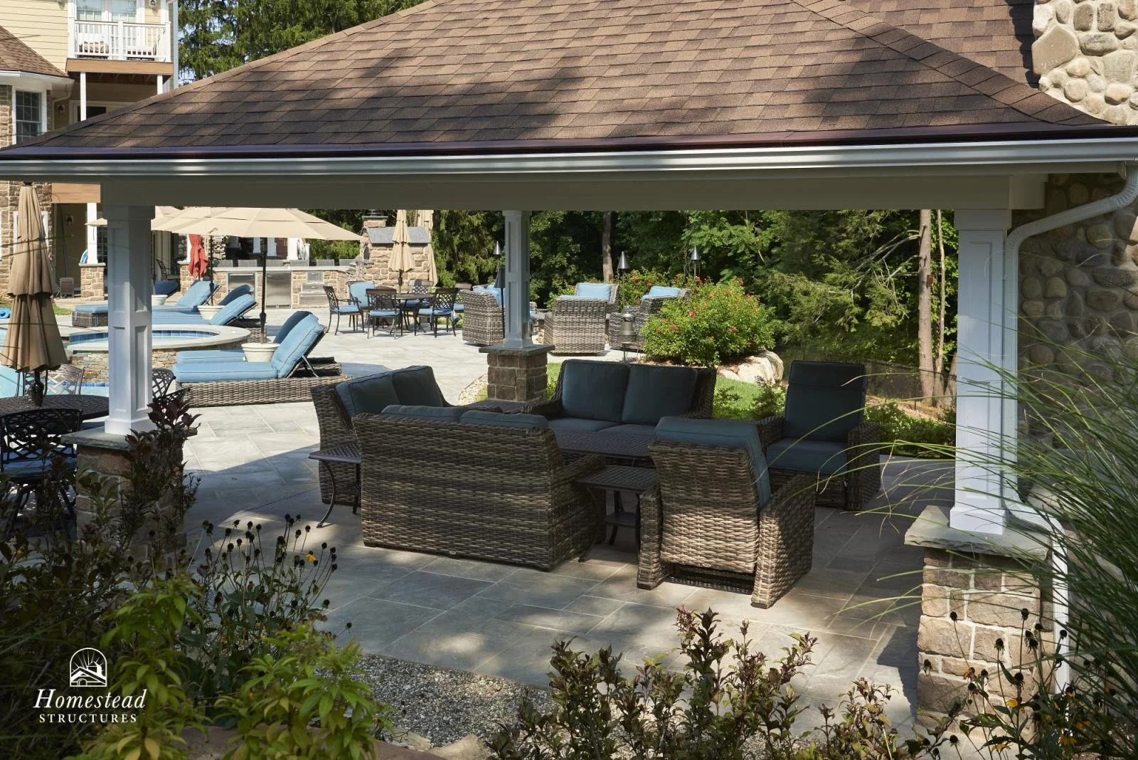 Outdoor patio area with wicker furniture, umbrellas, and lounge chairs under a covered structure with a shingled roof.