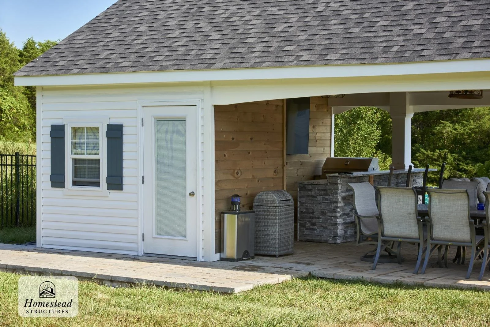 Backyard patio with an outdoor kitchen, dining table with chairs, white house wall with window and door, and a grassy yard.