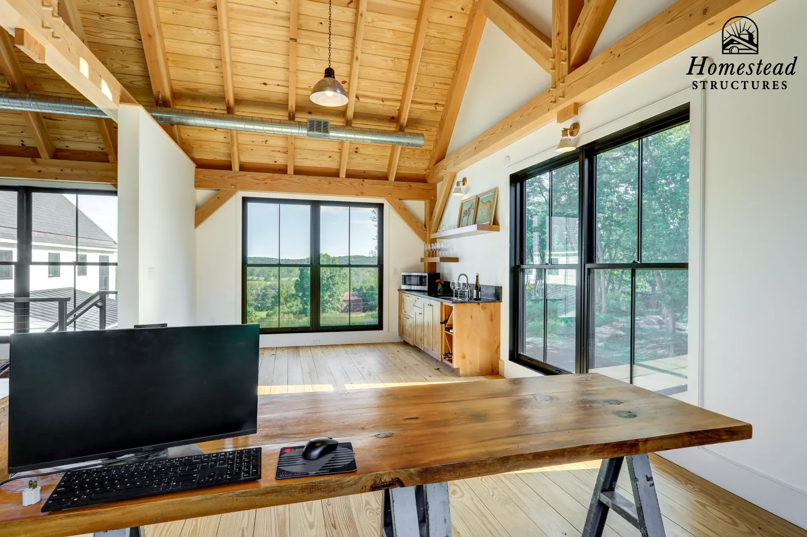 Interior of a modern wooden house with large windows, wooden ceiling beams, and a workspace with a computer on a wooden table.