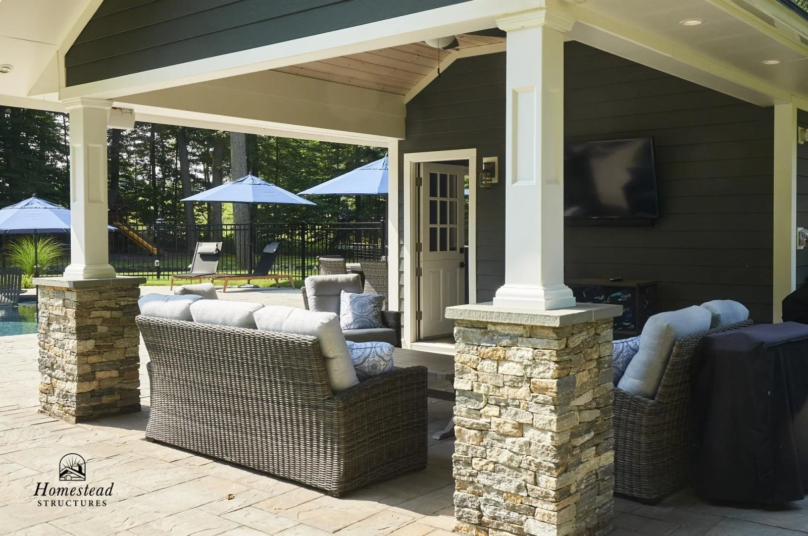 Outdoor patio with wicker seating, white cushions, stone pillars, a television mounted on a dark siding wall, and a backyard with umbrellas, lounge chairs, and trees.