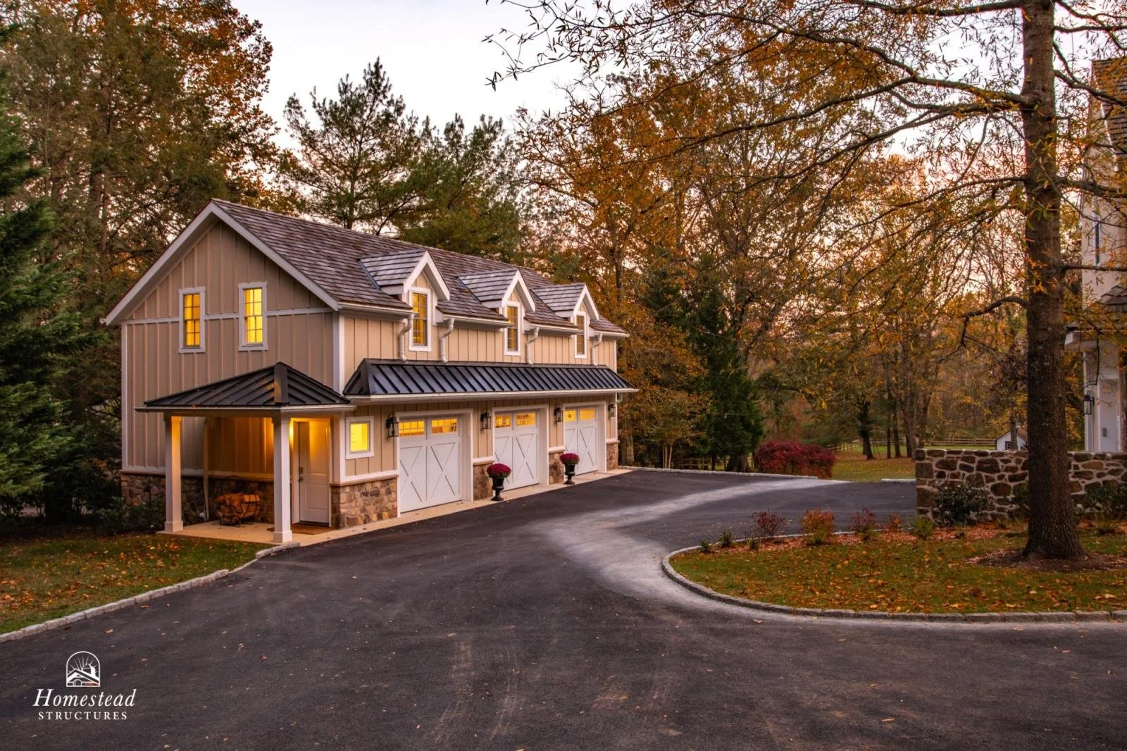 A two-story garage with a rustic design, beige siding, stone accents, three white barn doors, yellow-lit windows, and a black metal roof, surrounded by trees with fall foliage and a curved paved driveway at dusk.