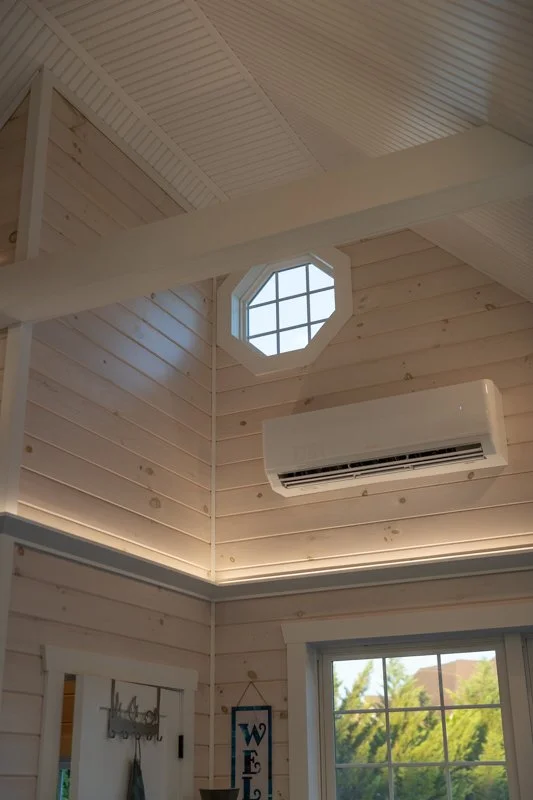 Interior of a wooden house with a high ceiling, octagonal window, air conditioning unit, and a window showing a green outdoor view.
