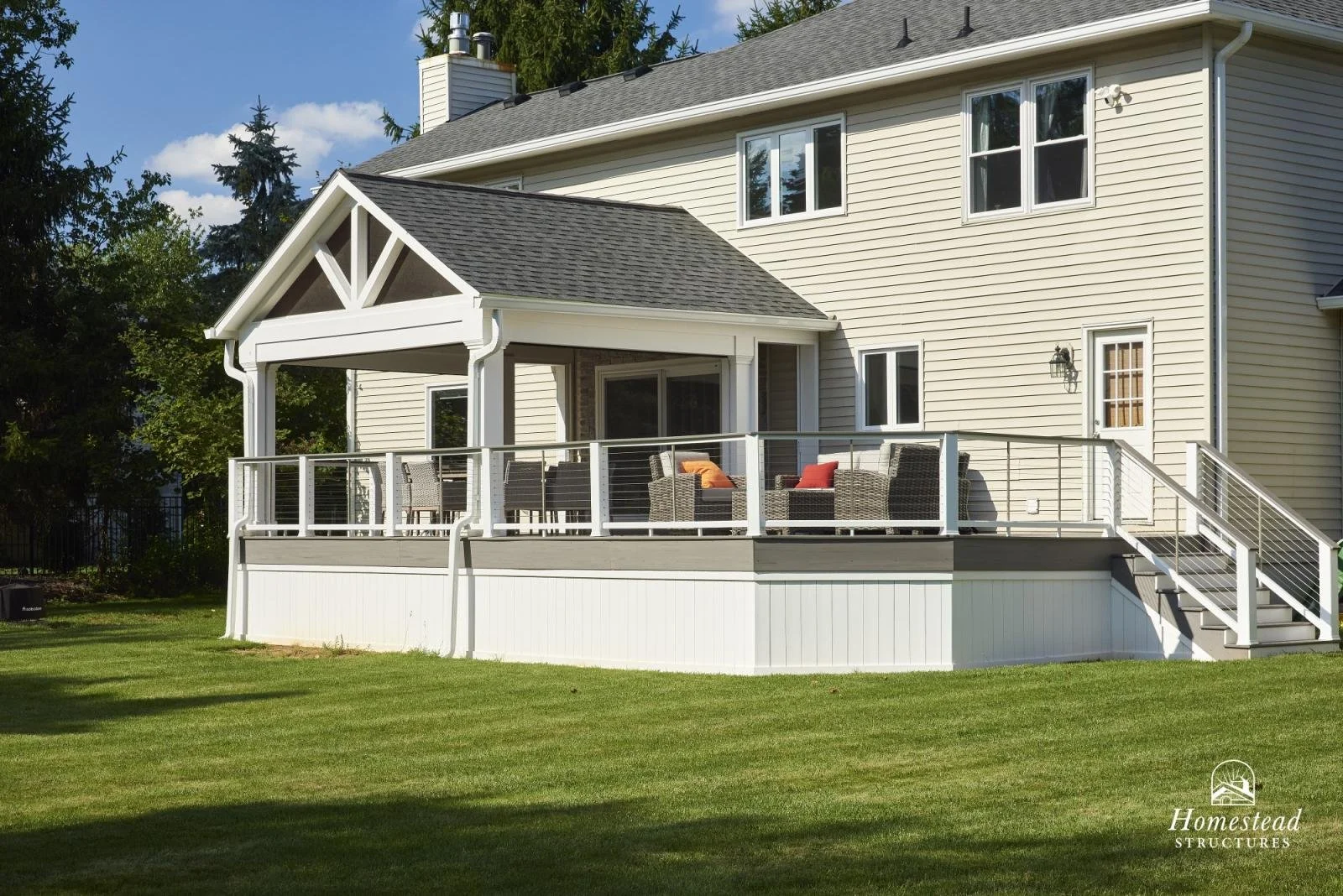 Back view of a two-story house with a large elevated deck featuring wicker furniture and colorful cushions, surrounded by a well-maintained lawn and trees, with clear sky overhead.