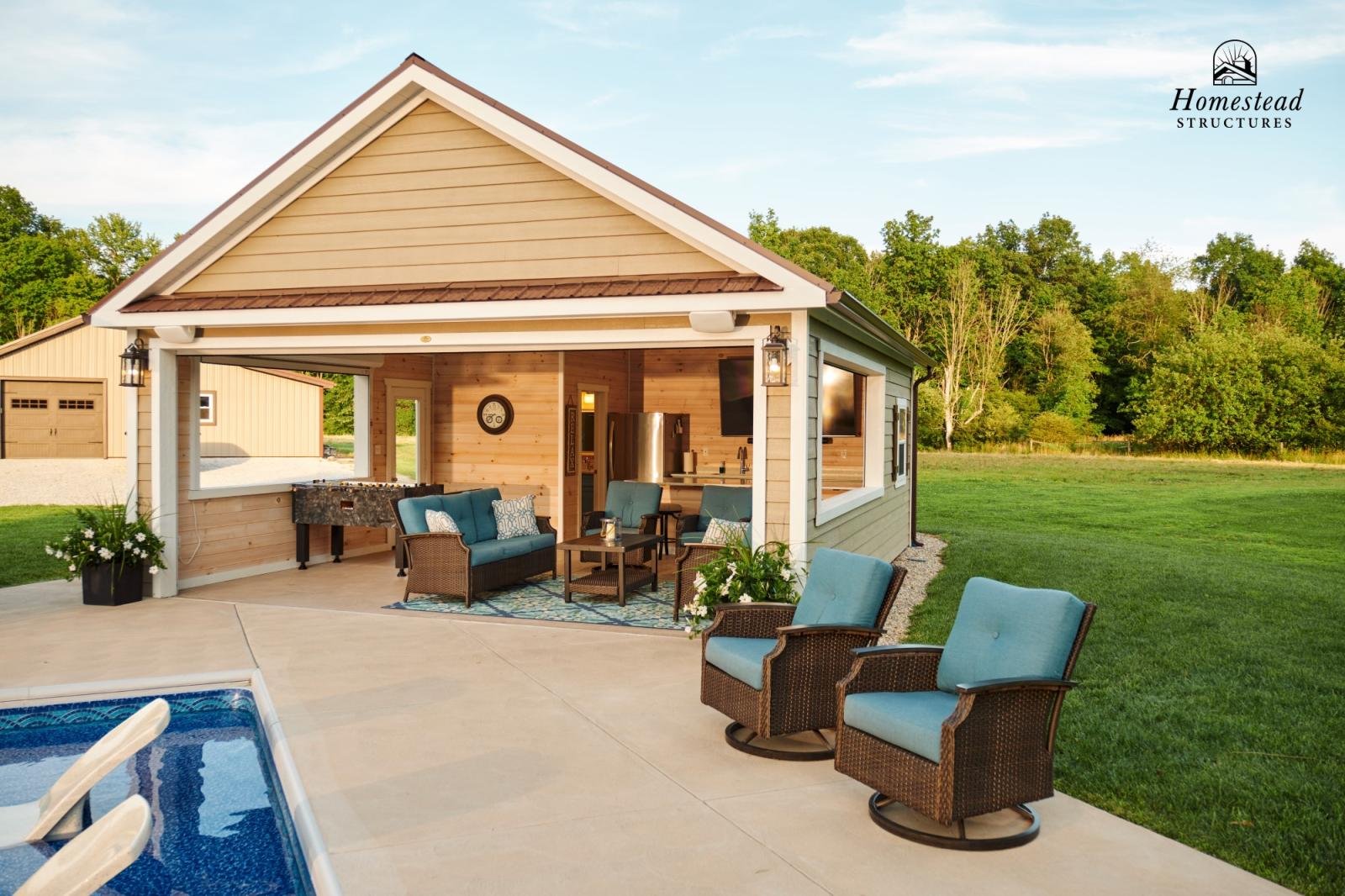 A backyard patio area with outdoor seating under a covered section, featuring a small sofa, armchairs, and a rug, with a pool in the foreground and a grassy lawn with trees in the background. The house has beige siding with wood accents and outdoor l