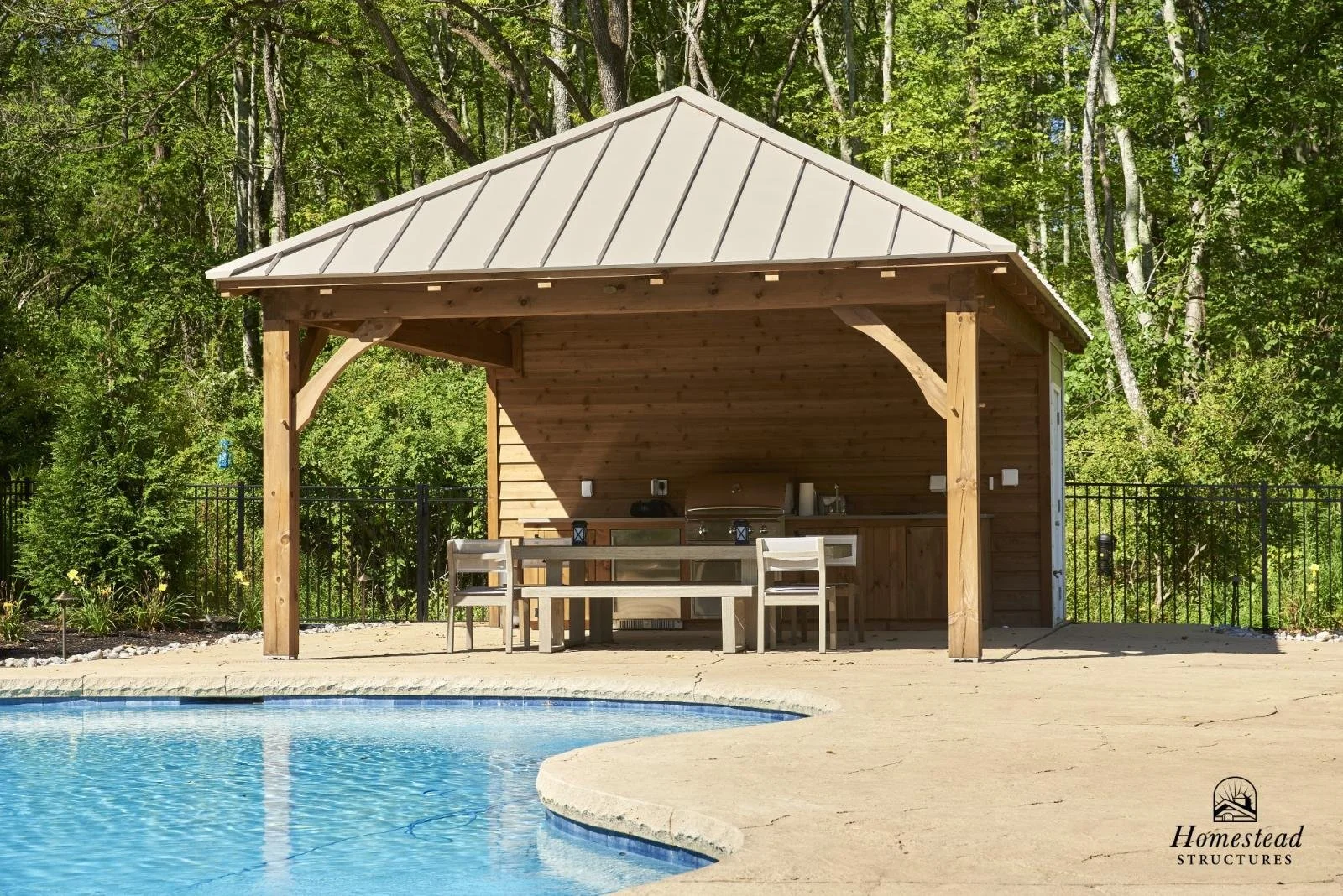 A backyard scene featuring a swimming pool with a concrete deck, and a wooden poolside cabana with a pitched metal roof, outdoor furniture, and a fenced wooded area in the background.