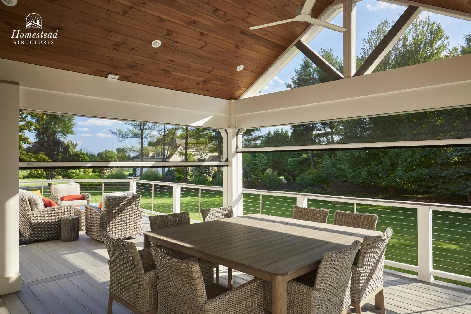 A covered outdoor patio with wicker chairs and a wooden dining table, overlooking a lush green yard with trees.