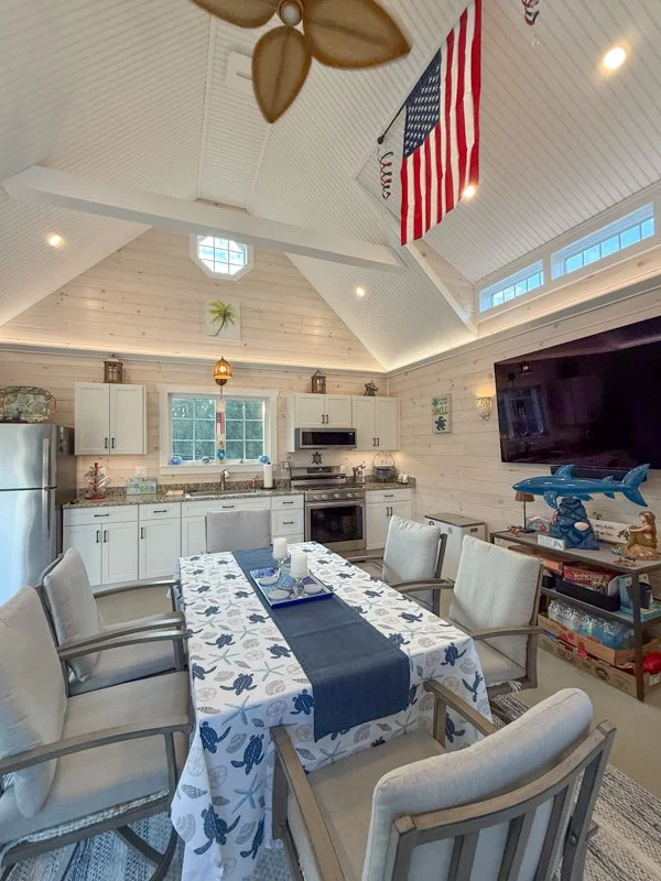 Beach-themed dining area with a white tablecloth featuring blue seashell and fish patterns, six beige upholstered chairs, a kitchen in the background with white cabinets and stainless steel appliances, and nautical decorations.