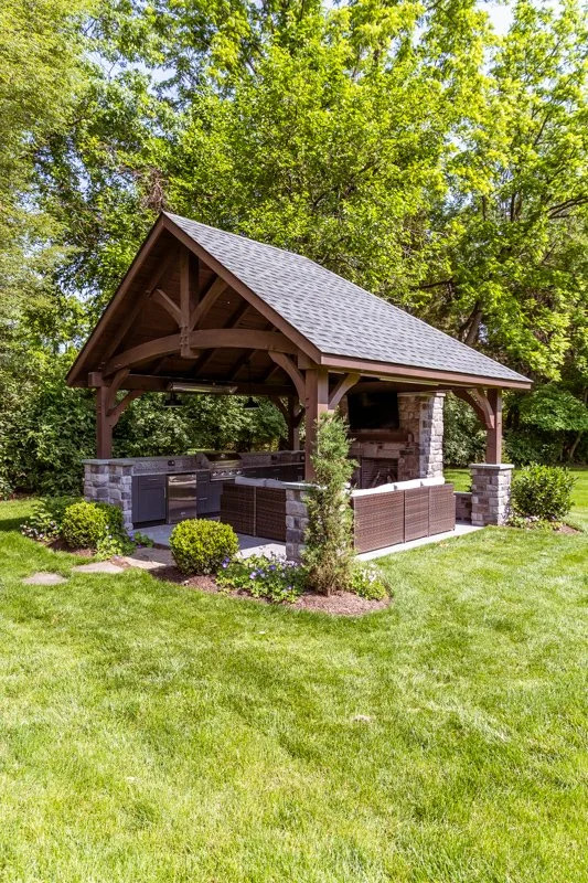 Covered outdoor grill area with stone pillars and a wooden roof, surrounded by a well-maintained lawn and green trees in the background.