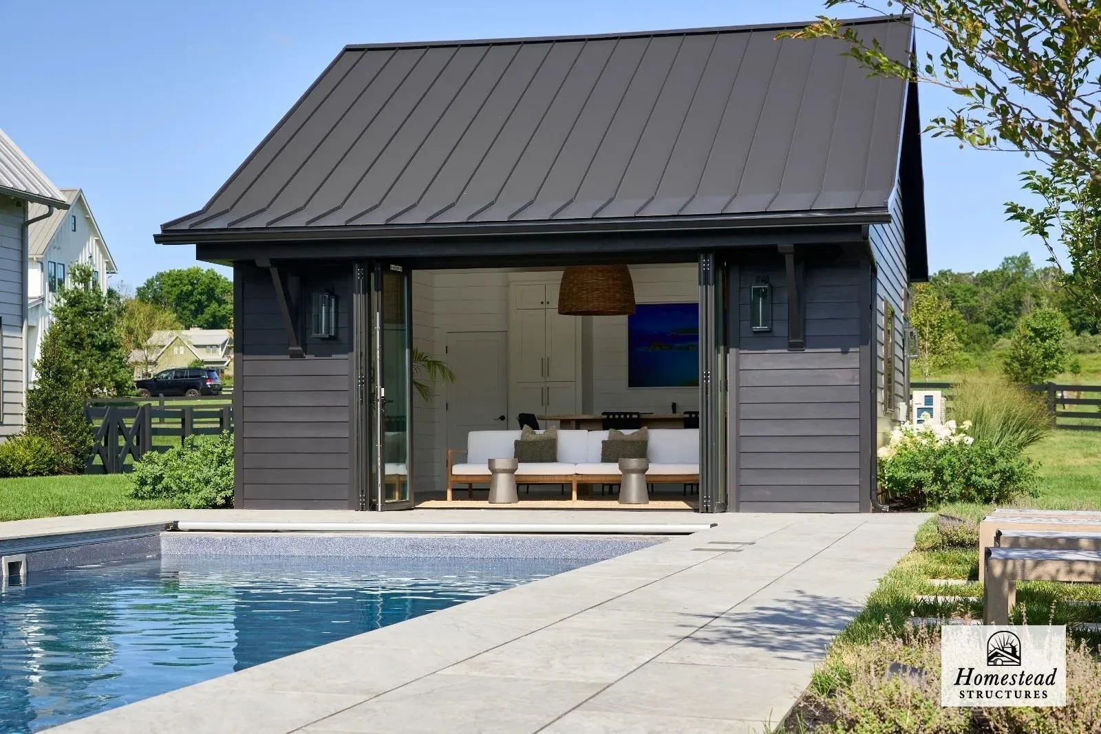 A black modern tiny house with sliding glass doors, indoor seating, a flat-screen TV, and a hanging pendant light. It is situated beside a swimming pool and surrounded by a green lawn with trees and neighboring houses in the background.