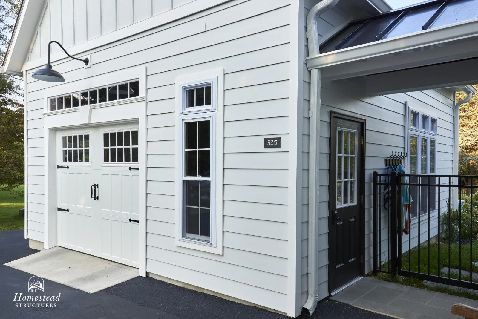 Exterior view of a modern white house with a garage door, a small black door, and several windows, including a large window near the garage and a narrow window beside the black door. There is a house number '325' on the wall, a black outdoor light fi