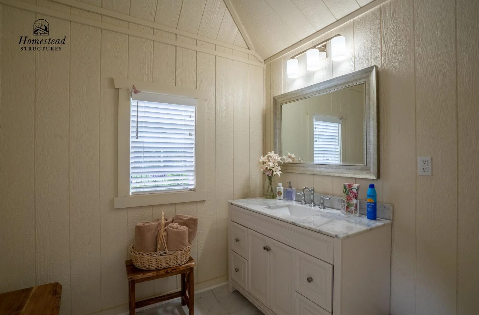 Small bathroom with beige wooden paneled walls, white vanity with marble countertop, large mirror, two-lamp light fixture, window with blinds, pink lilies in a vase, toiletries, towel basket, and natural lighting.