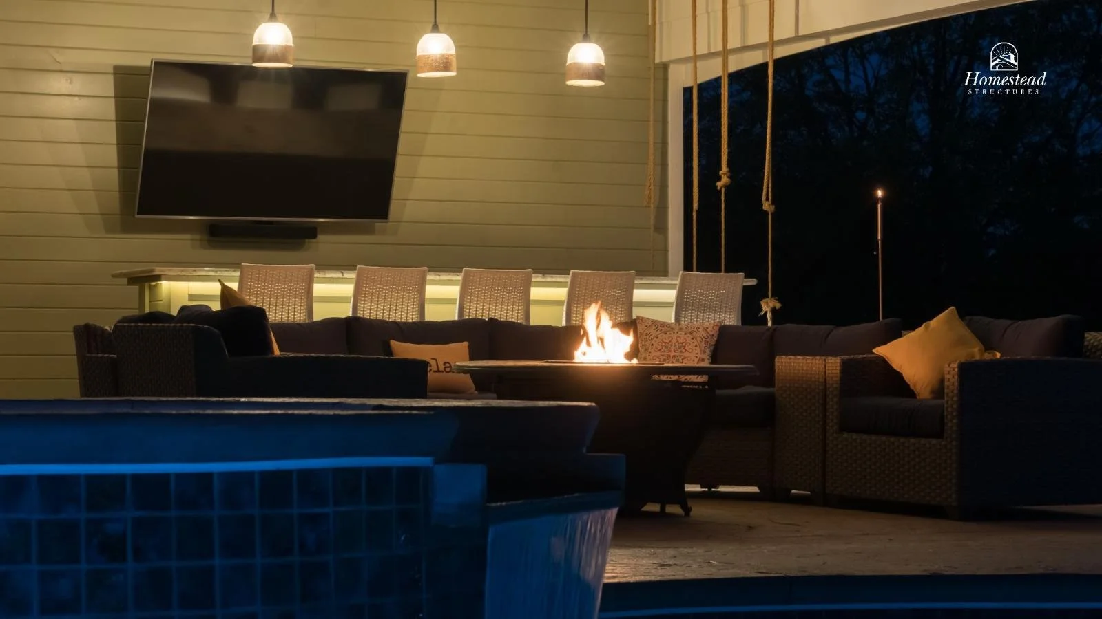 Outdoor patio with a fire pit, sectional sofa with pillows, bar area with chairs, and a mounted flat-screen TV on a wooden wall, nighttime setting.
