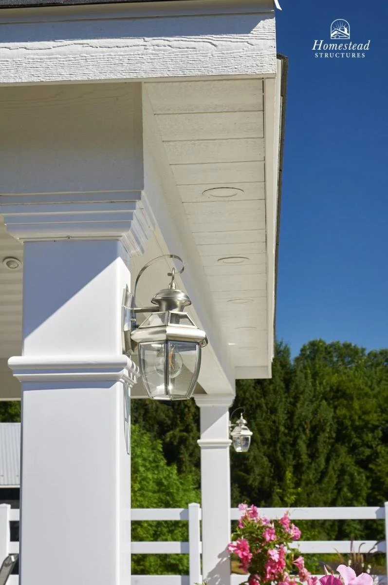 White porch columns with lantern-style light fixtures, pink flowers, white picket fence, and lush green trees in the background under a clear blue sky.