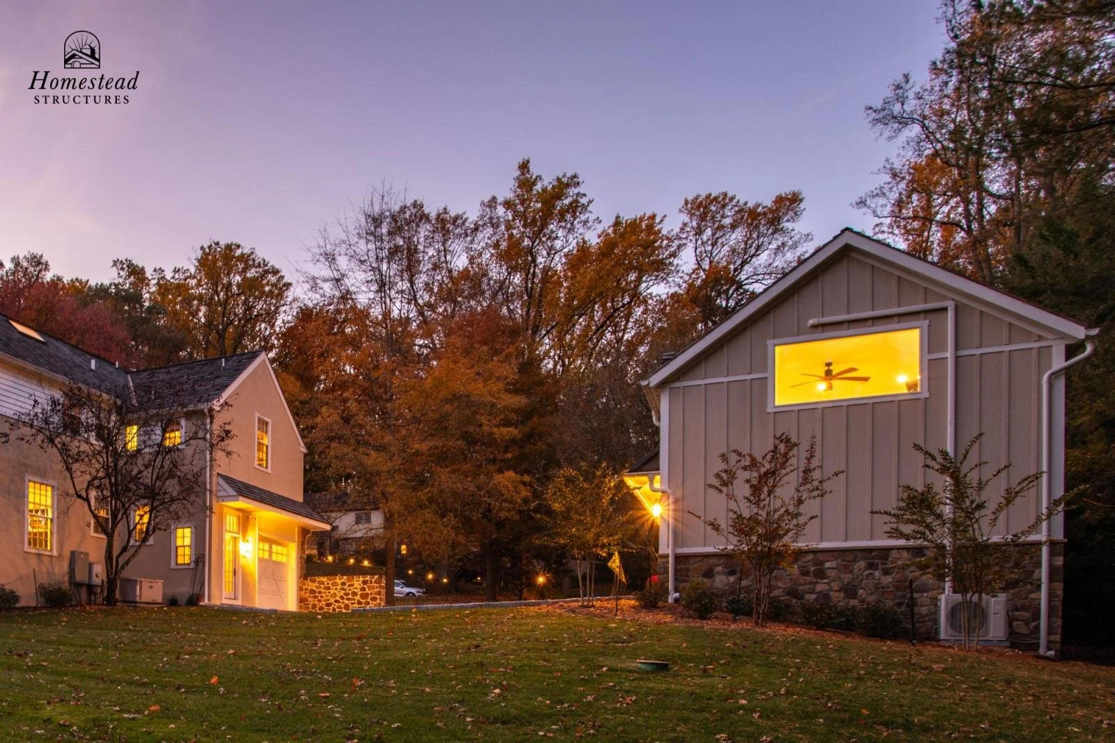 Two houses illuminated in the evening with autumn trees in the background. The house on the left has warm yellow lights in the windows, and the house on the right has a bright yellow light in a window. The sky is dusky, and there are leaves on the gr