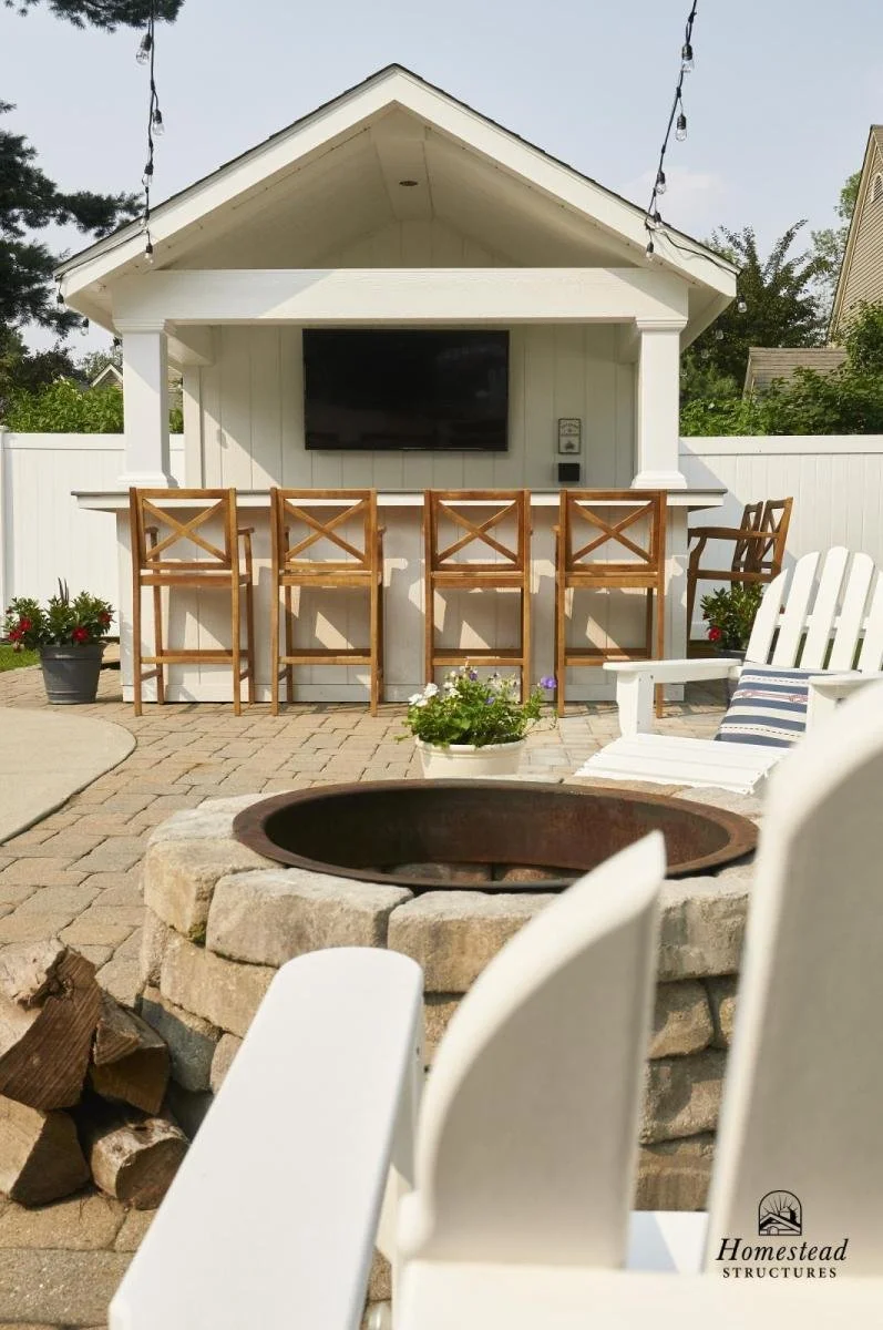 Outdoor backyard entertainment area featuring a white bar with four wooden chairs, a mounted flat-screen TV, string lights, a firepit with a stone surround, a white Adirondack chair with a striped cushion, potted plants, and a privacy fence.
