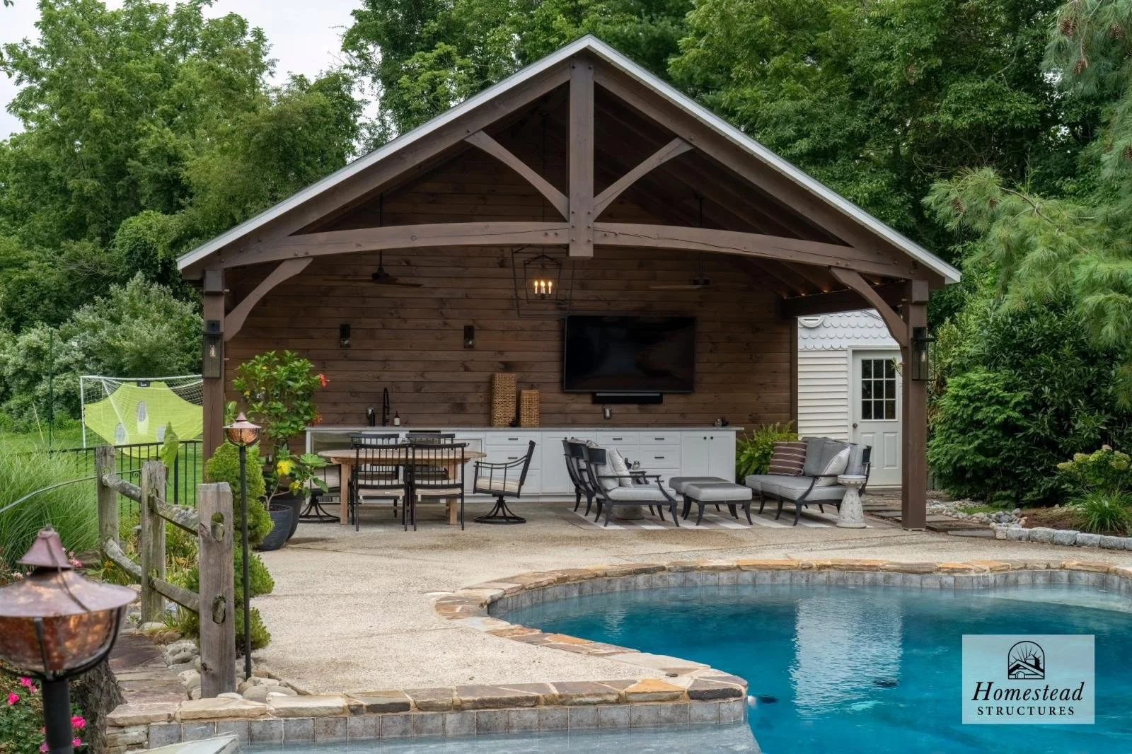 An outdoor living space with a wooden pavilion housing a television, seating area, and outdoor kitchen, adjacent to a swimming pool surrounded by lush green trees and landscaping.