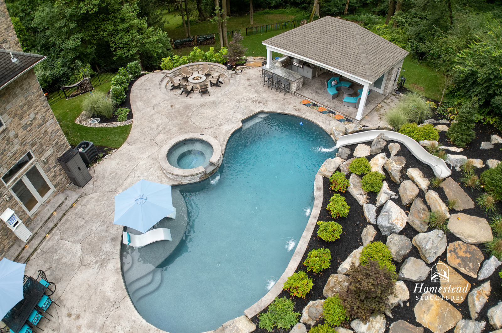 A backyard with a swimming pool, hot tub, pool slide, hot tub, patio with umbrellas, outdoor furniture, fire pit surrounded by chairs, and a covered outdoor kitchen area, all surrounded by lush green trees.