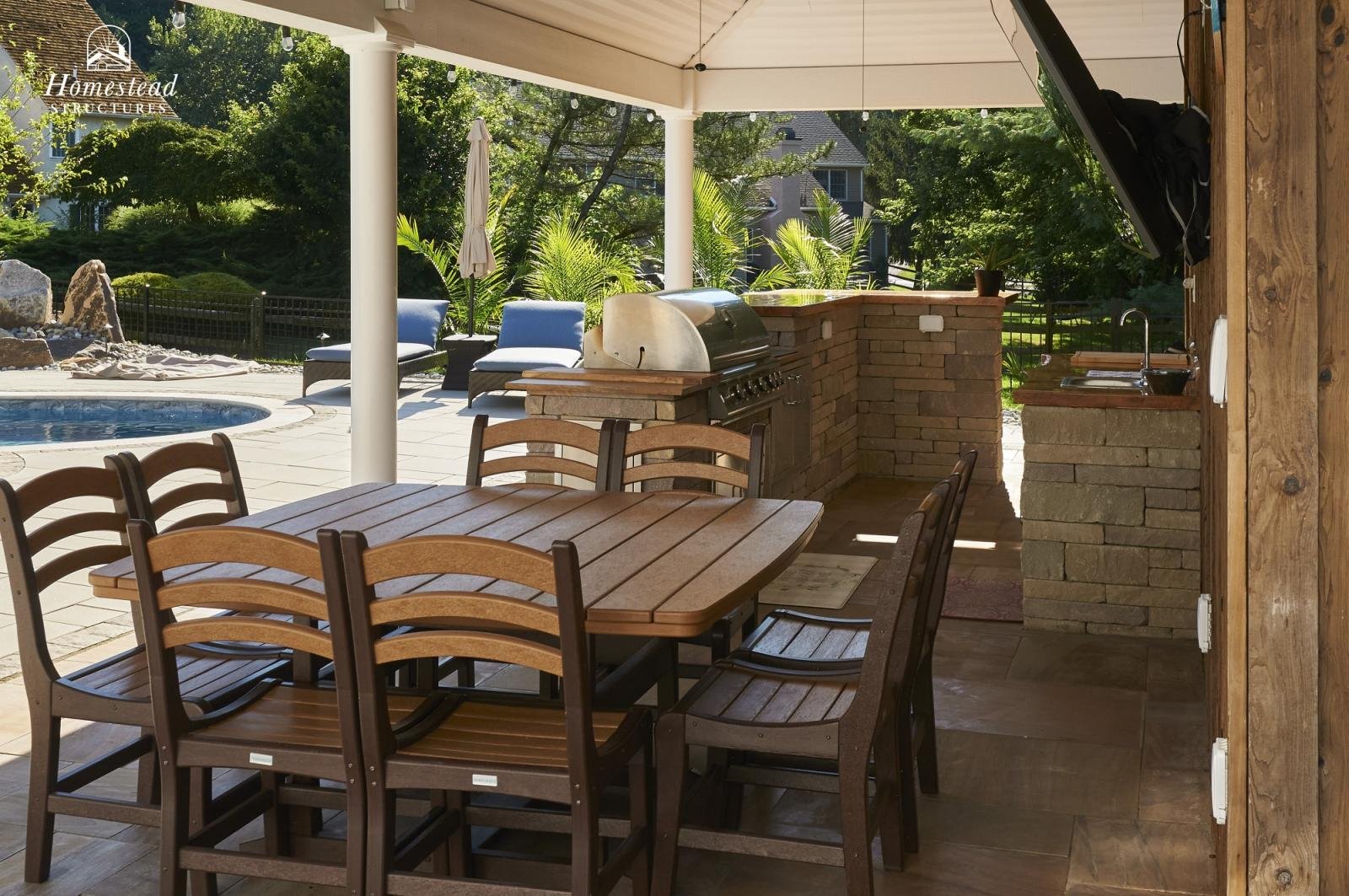 Outdoor patio with wooden dining table and chairs, brick barbecue grill, outdoor kitchen sink, and pool with lounge chairs surrounded by trees and greenery