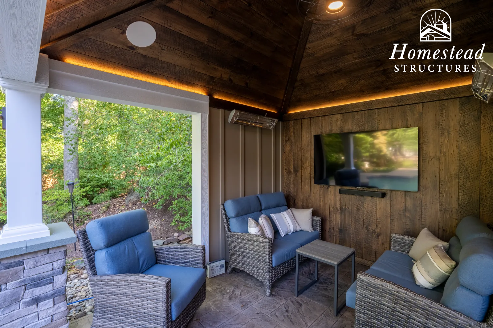 Outdoor living space with wicker furniture with blue cushions, a wall-mounted TV, and a wooden ceiling with lighting, overlooking a yard with trees.