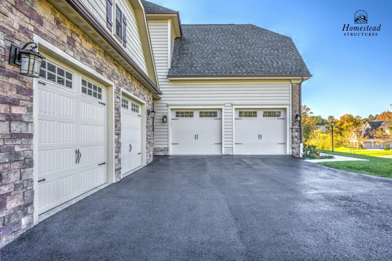 A modern residential driveway with four white garage doors, two with windows at the top, flanked by stone and siding exterior walls, with yard and trees in the background and a clear sky.