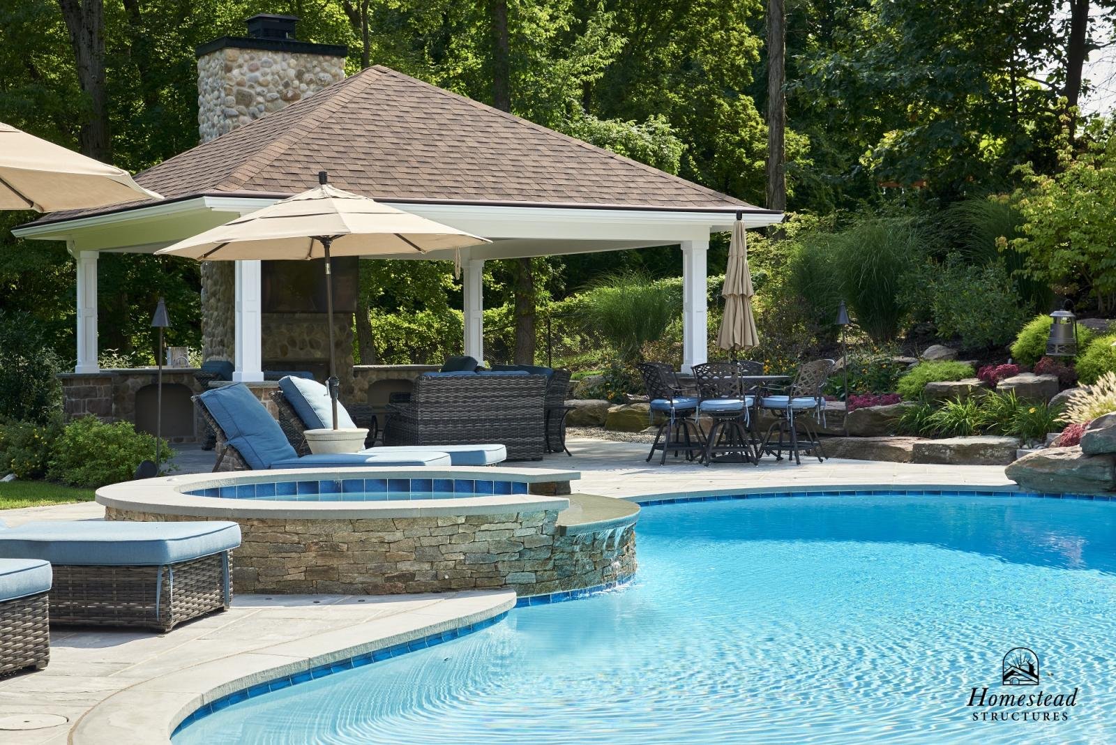 A backyard with a pool, spa, outdoor seating and dining area, and a shaded pavilion surrounded by greenery.