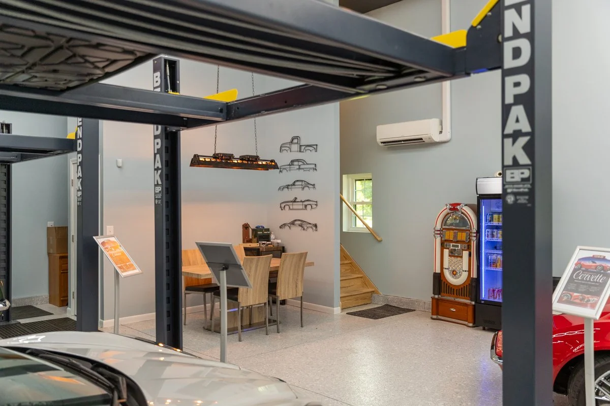 Inside a car museum showroom with car lifts, seating area, vintage jukebox, and display signs for classic cars.